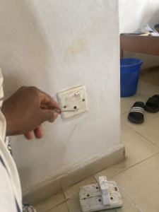 student fixing a hostel wall socket with a small screwdriver, extension box on floor nearby