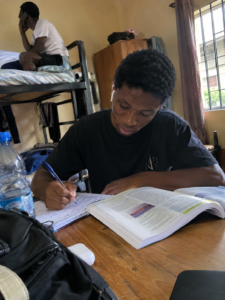 Nigerian university student studying at a hostel desk with earbuds in while roommate talks in the background